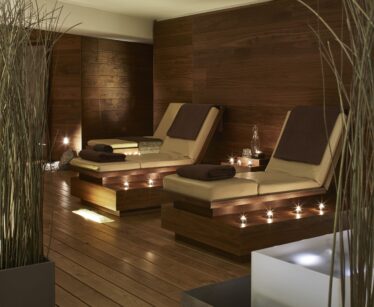 A modern spa relaxation room featuring two padded loungers with brown towels, dim lighting, and decorative tall grass.