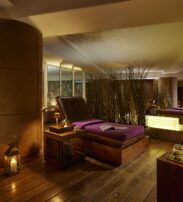 A dimly lit spa room with wooden floors, lounge chairs with purple cushions, candles, and decorative screens creating a relaxing atmosphere.