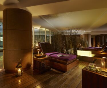 A dimly lit spa room with wooden floors, lounge chairs with purple cushions, candles, and decorative screens creating a relaxing atmosphere.