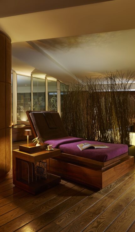 A dimly lit spa room with wooden floors, lounge chairs with purple cushions, candles, and decorative screens creating a relaxing atmosphere.