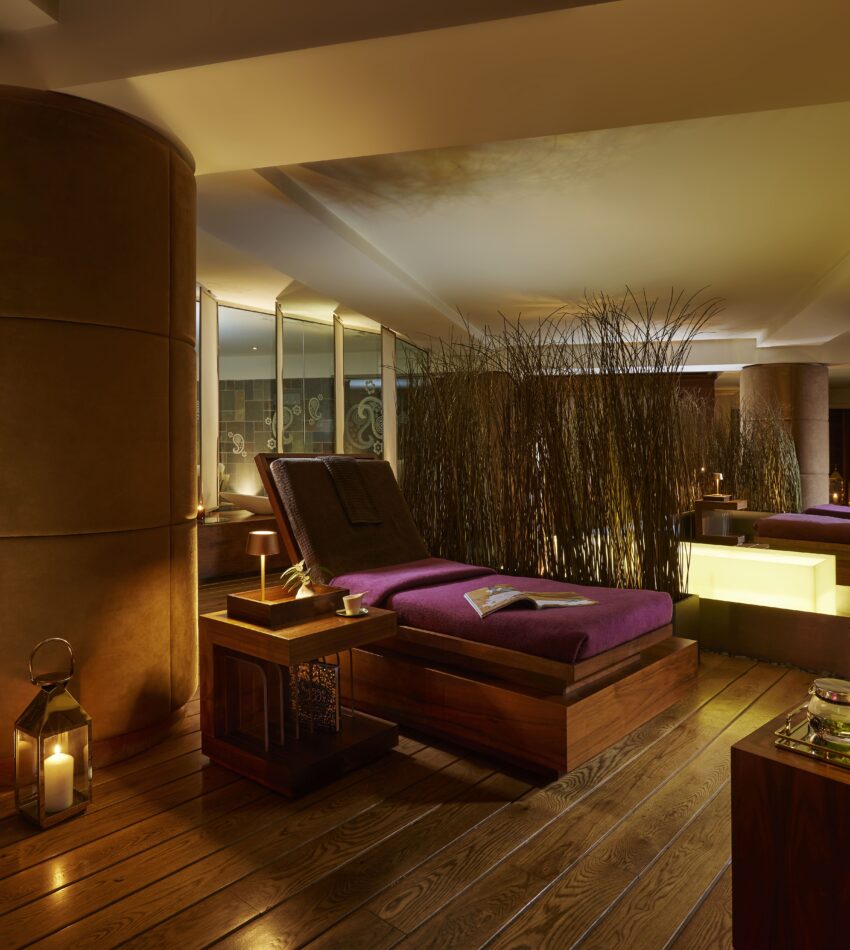A dimly lit spa room with wooden floors, lounge chairs with purple cushions, candles, and decorative screens creating a relaxing atmosphere.