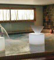 A person relaxes in an indoor spa pool with water features and stone wall tiles, next to illuminated structures and a window with decorative reeds.