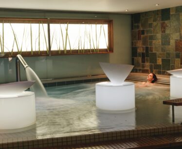 A person relaxes in an indoor spa pool with water features and stone wall tiles, next to illuminated structures and a window with decorative reeds.
