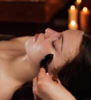 A woman lying down receives a facial treatment with a jade roller in a dimly lit room; candles are visible in the background.