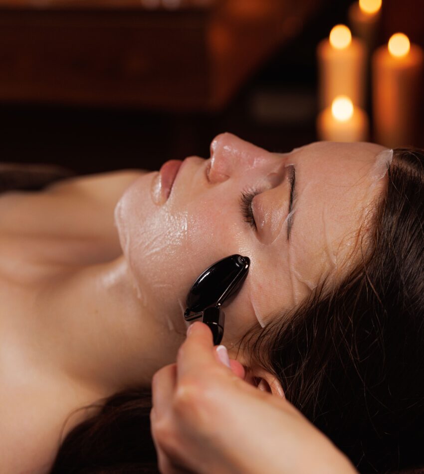 A woman lying down receives a facial treatment with a jade roller in a dimly lit room; candles are visible in the background.