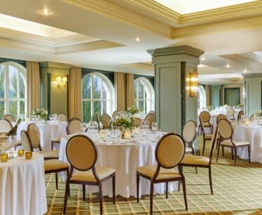 Elegant banquet hall with round tables covered in white tablecloths, set for an event, with formal chairs and floral centrepieces near large arched windows.