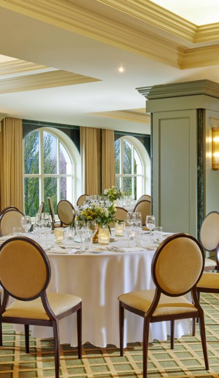 Elegant banquet hall with round tables covered in white tablecloths, set for an event, with formal chairs and floral centrepieces near large arched windows.