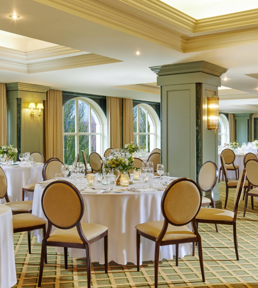 Elegant banquet hall with round tables covered in white tablecloths, set for an event, with formal chairs and floral centrepieces near large arched windows.