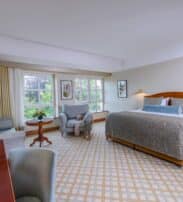 Spacious hotel room with a king-size bed, armchairs, sofa, desk, large windows with curtains, and neutral-coloured decor.