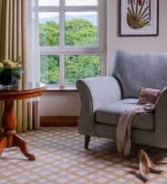 A grey armchair with a handbag and scarf draped on it, beside a round wooden table with a flower vase, in a room with large windows and beige curtains.