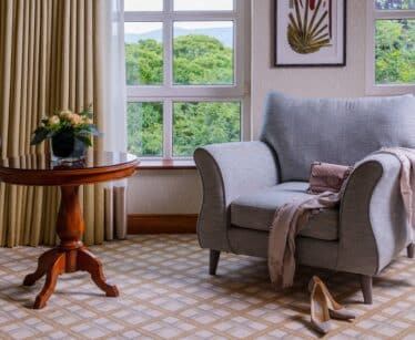 A grey armchair with a handbag and scarf draped on it, beside a round wooden table with a flower vase, in a room with large windows and beige curtains.