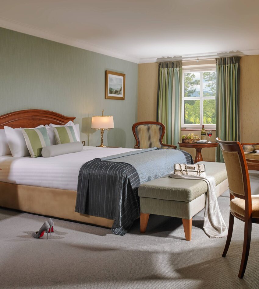 A neatly arranged hotel room with a large bed, bedside tables, armchairs, a writing desk, and a window with green curtains letting in natural light.