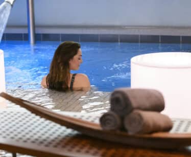 A woman relaxes in an indoor pool near a water feature, with rolled towels and two illuminated lamps in the foreground.