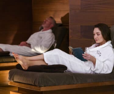 Two people wearing white robes relax on loungers; one is reading a book while the other has eyes closed in a dimly lit spa setting.