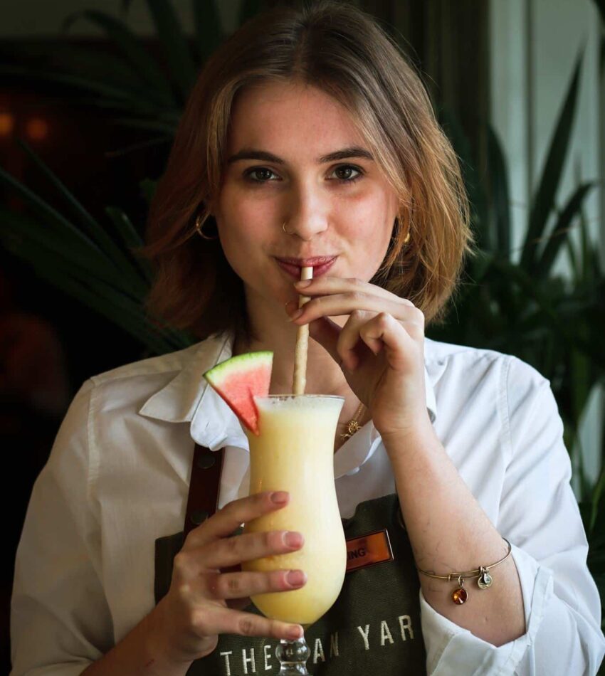 A person wearing a white shirt and apron drinks a yellow smoothie with a straw from a tall glass garnished with a watermelon slice.