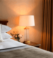A neatly made bed with white linen and decorative cushions next to a wooden bedside table with a lamp, a small flower arrangement, and a window with brown curtains.