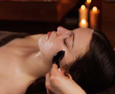 A person receives a facial massage with a black jade roller whilst lying down with eyes closed; lit candles are blurred in the background.