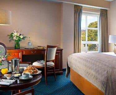 A hotel room with a made bed, desk, chair, lamp, flowers, and a round table set with breakfast items near a window with curtains.