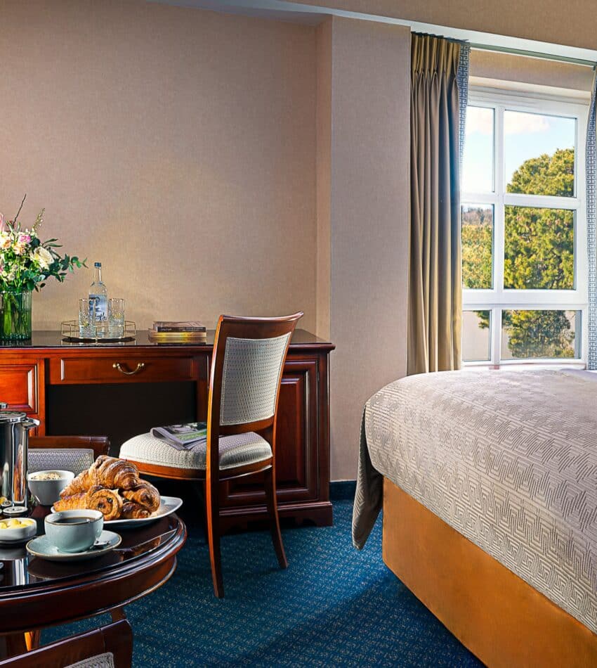 A hotel room with a made bed, desk, chair, lamp, flowers, and a round table set with breakfast items near a window with curtains.