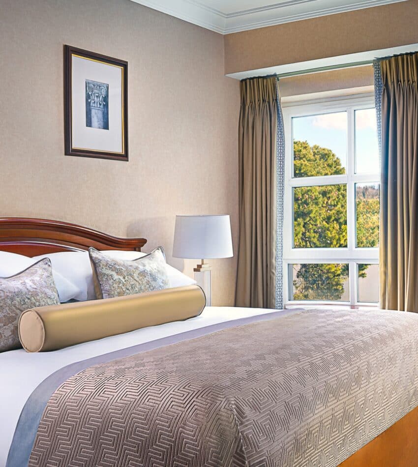 A neatly made hotel room bed with decorative cushions and a bolster, beside a window with curtains, two framed artworks, a lamp, and a wooden desk and chair.