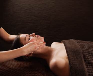 A person lying on a massage table receives a facial massage from a therapist, covered by a brown towel, against a dark background.