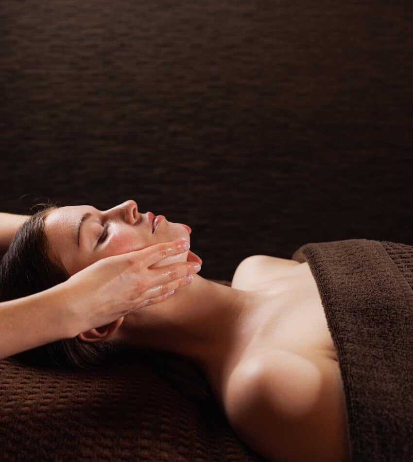 A person lying on a massage table receives a facial massage from a therapist, covered by a brown towel, against a dark background.
