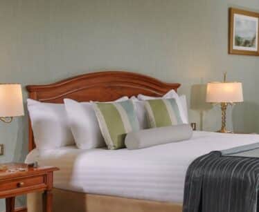 A neatly made hotel bed with white linen, striped accent cushions, a wooden headboard, two bedside tables with lamps, and a framed picture on the wall.