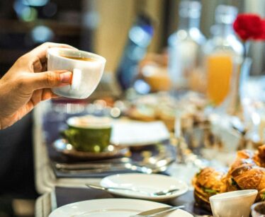 A person holds a white cup of coffee at a breakfast table set with plates, pastries, juice, and a red flower in a vase.