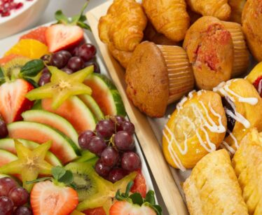 A platter of sliced fresh fruit including watermelon, strawberries, grapes, and kiwi next to an assortment of muffins and pastries with icing.