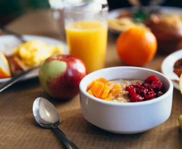 A breakfast table with a bowl of porridge topped with fruit, an apple, orange juice, fruit, a muffin, and plates of assorted breakfast foods.