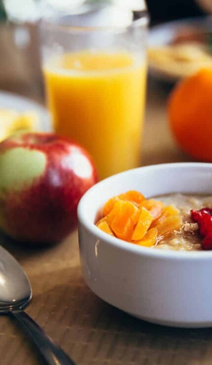 A breakfast table with a bowl of porridge topped with fruit, an apple, orange juice, fruit, a muffin, and plates of assorted breakfast foods.