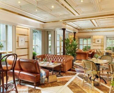 Elegant lounge with brown leather sofas, marble floors, plants, and large windows letting in natural light; tables are set for dining.