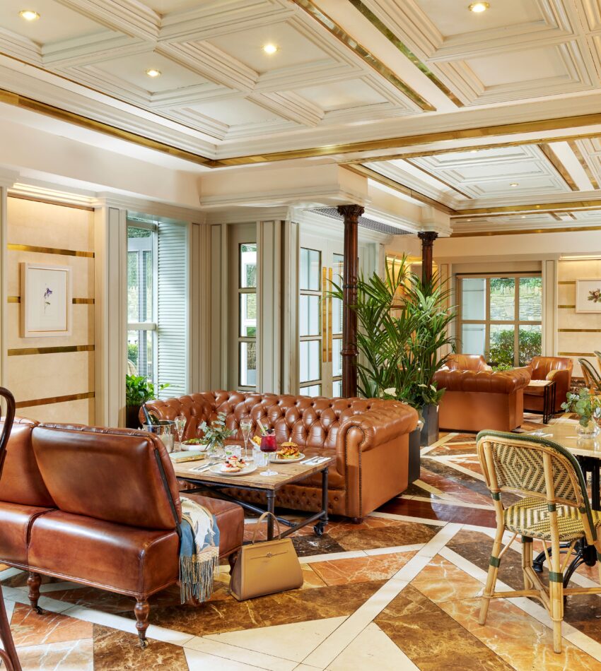 Elegant lounge with brown leather sofas, marble floors, plants, and large windows letting in natural light; tables are set for dining.