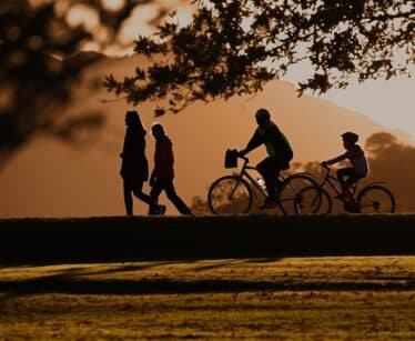 Silhouettes of two people walking and two people riding bicycles on a grassy path at sunset, with trees and mountains in the background.