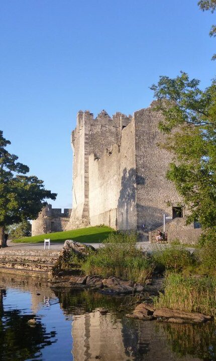 A stone castle stands next to a lake, partially surrounded by trees and greenery, with its reflection visible in the calm water under a clear blue sky.
