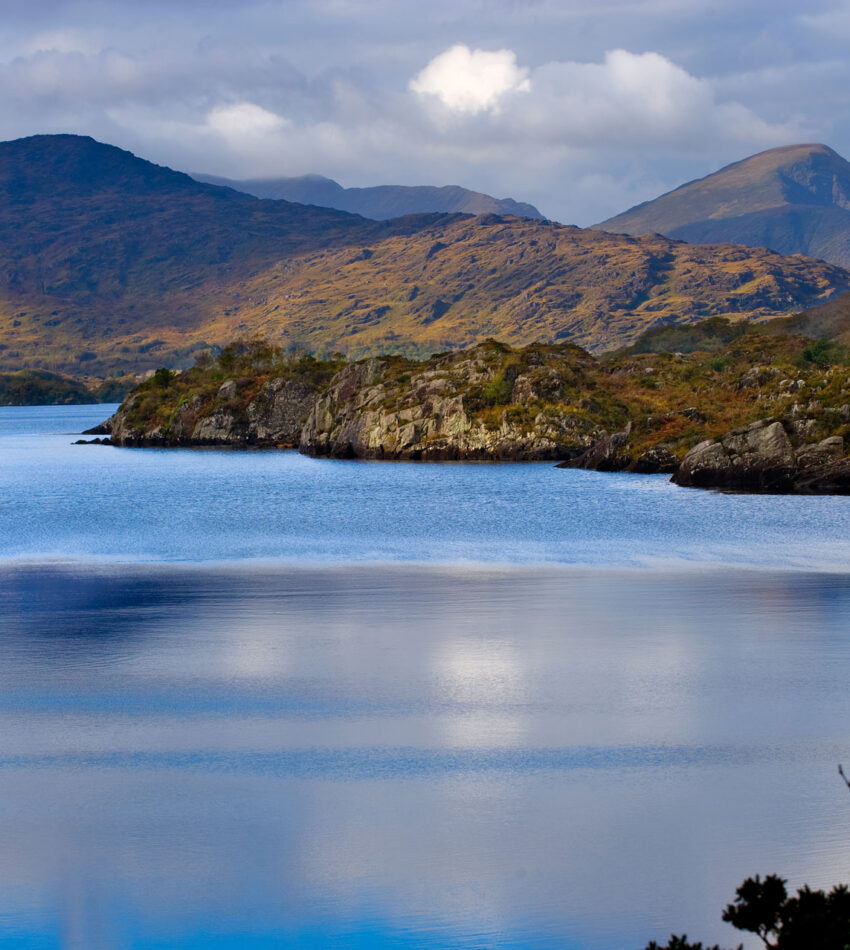 A calm lake bordered by rocky hills and mountains under a cloudy sky, with reflections visible on the water’s surface.