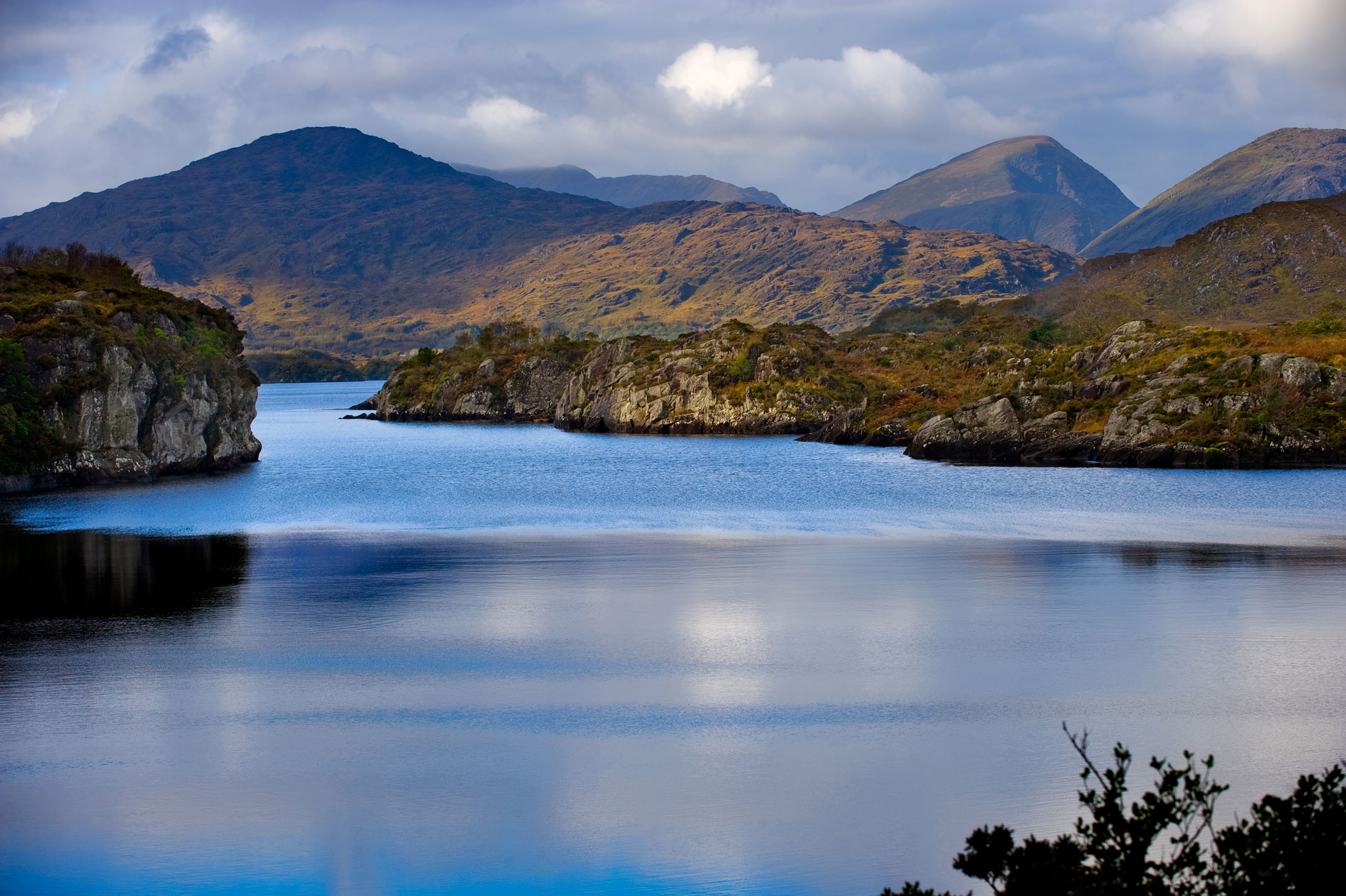 A calm lake bordered by rocky hills and mountains under a cloudy sky, with reflections visible on the water’s surface.