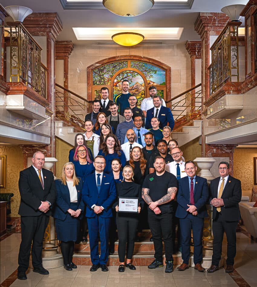 A group of formally dressed people pose on a marble staircase in a hotel reception, with one woman holding a framed certificate at the front.