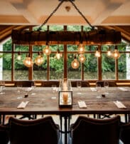 A rustic dining room features a large wooden table set for eight, surrounded by leather chairs, beneath hanging Edison bulbs and in front of tall windows with a green outdoor view.