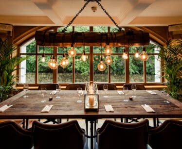 A rustic dining room features a large wooden table set for eight, surrounded by leather chairs, beneath hanging Edison bulbs and in front of tall windows with a green outdoor view.