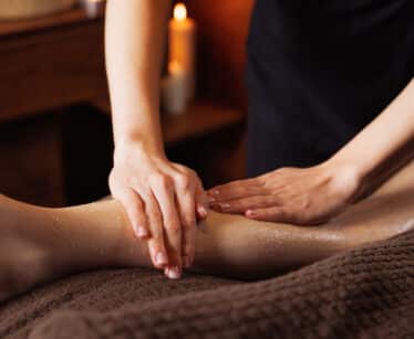 A person massages another person's lower leg with exfoliating scrub in a spa setting, with a lit candle in the background.