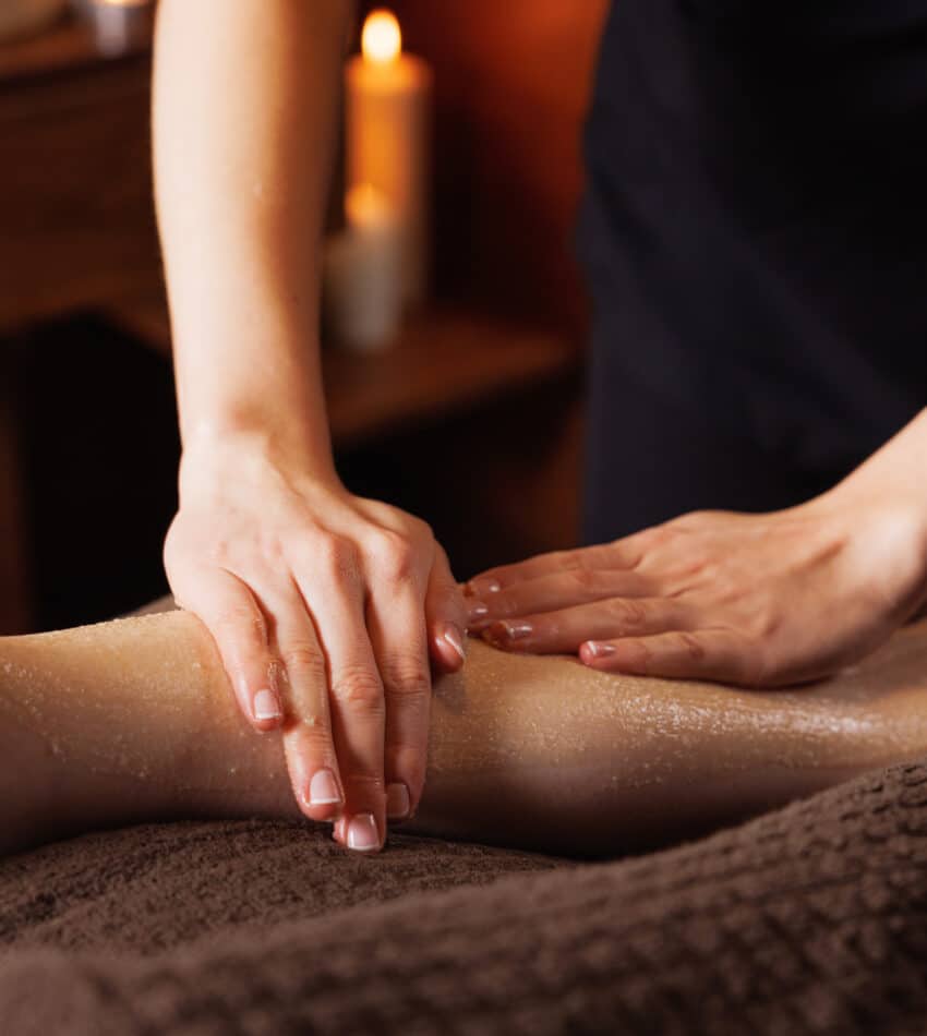 A person massages another person's lower leg with exfoliating scrub in a spa setting, with a lit candle in the background.