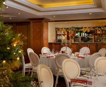 A decorated dining area with round tables set for a meal, white chairs, a Christmas tree in the foreground, and a bar with festive garland in the background.
