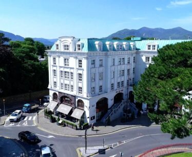 A large white hotel with a green roof stands at a curved junction, surrounded by trees and mountains under a clear blue sky. Cars are parked and driving on the street.