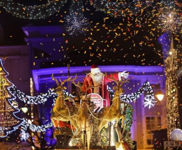 Father Christmas waves from a sleigh with reindeer during a nighttime holiday parade, surrounded by festive lights, decorations, and falling confetti.