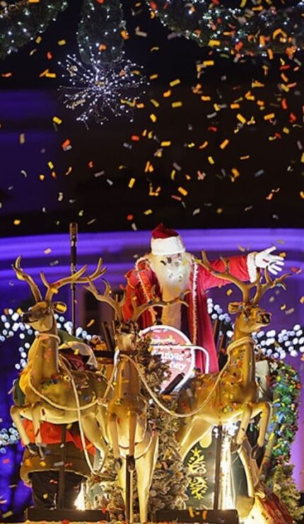 Father Christmas waves from a sleigh with reindeer during a nighttime holiday parade, surrounded by festive lights, decorations, and falling confetti.