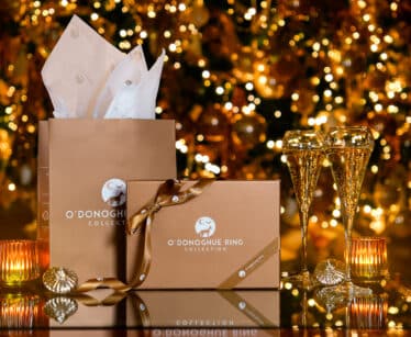 Gift boxes and two champagne glasses are arranged on a reflective surface with a decorated, lit Christmas tree in the background.