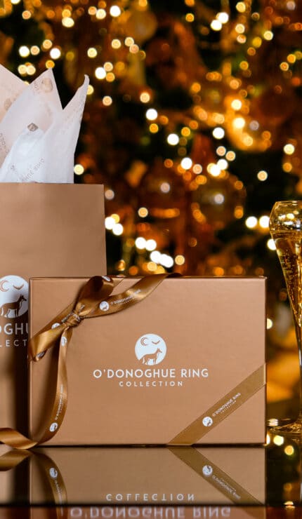 Gift boxes and two champagne glasses are arranged on a reflective surface with a decorated, lit Christmas tree in the background.