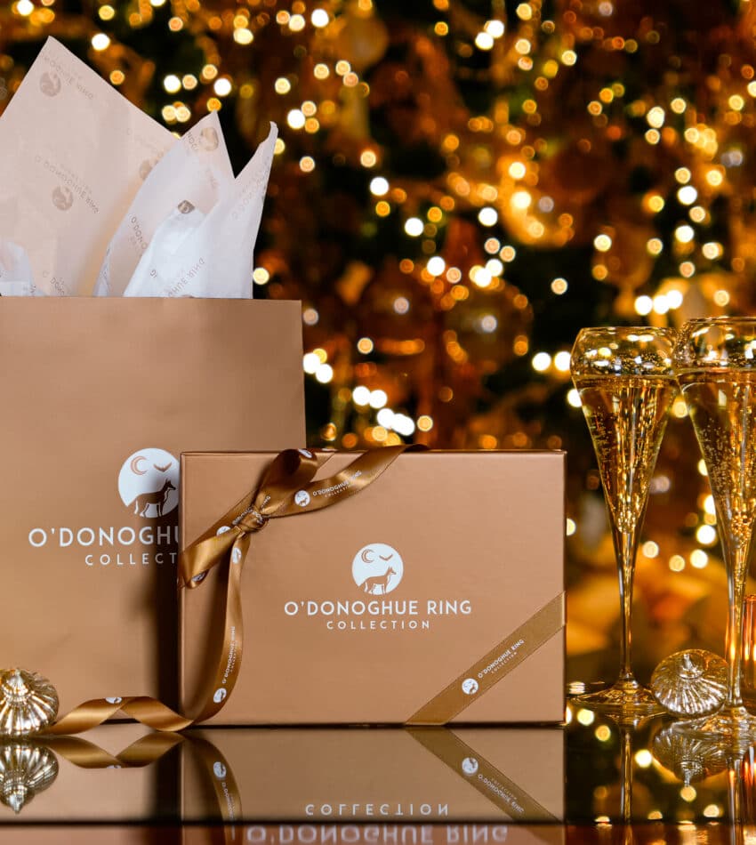 Gift boxes and two champagne glasses are arranged on a reflective surface with a decorated, lit Christmas tree in the background.