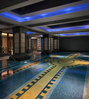 Indoor swimming pool with tiled mosaic floor, illuminated by blue recessed ceiling lights and surrounded by black pillars and walls. Steps lead into the water on both sides.